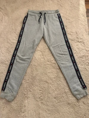 Tommy Hilfiger mjukisbyxor - Snygga grå mjukisbyxor från Tommy Hilfiger med svarta dragsnören i midjan och ribbade muddar vid bensluten. Sidorna har en cool logga med texten 'TOMMY HILFIGER' och klassiska färgdetaljer. Perfekta för chill dagar och streetwear vibes.
