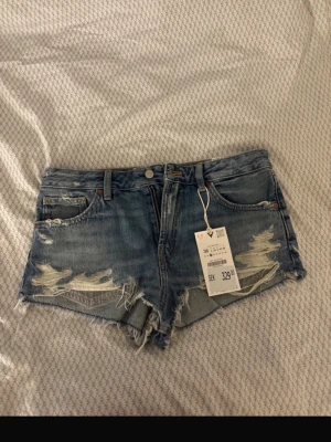 Jeansshorts Zara - Säljer ett par blå jeansshorts med slitna detaljer och fransig kant. Shortsen har fem fickor, bälteshällor och knappgylf. Perfekta för sommaren och ger en avslappnad vibe till din outfit. Pris går att diskutera 