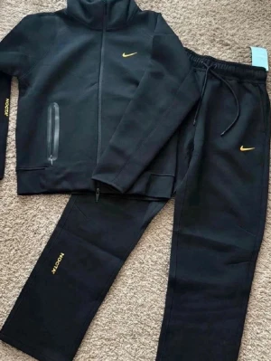 Svart Nike NOCTA träningsset - Säljer ett svart träningsset från Nike NOCTA med gul logga. Setet består av en zip-jacka med hög krage och fickor samt matchande byxor med dragsko i midjan. Både jacka och byxor har NOCTA-tryck i gult. Perfekt för gymmet eller chill.