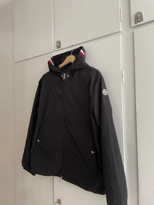 Svart vindjacka från Moncler - Snygg svart vindjacka från Moncler med huva och dragkedja framtill. Jackan har Moncler-logga på ärmen och detaljer i rött, vitt och blått på huvan. Två fickor med tryckknappar och justerbar nederkant. Perfekt för blåsiga dagar.