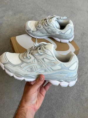 Vita och grå ASICS sneakers - Säljer ett par fräscha vita och ljusgrå ASICS sneakers med klassisk mesh och syntetpaneler. Skorna har en chunky sula och ikoniska ASICS-stripes på sidan. Perfekta för dig som gillar sportig och clean stil.