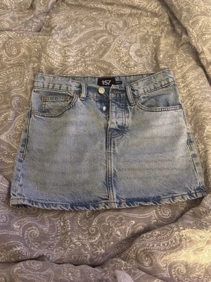 Ljusblå jeanskjol från 157 - Snygg ljusblå jeanskjol från 157 med klassisk femficksdesign, bälteshällor och knappstängning framtill. Kjolen är kort och har en rak passform, perfekt till många olika stilar.