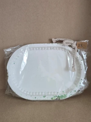 Vit oval dekoration med rep - Vit oval dekoration i gipsmed upphöjd kant och små detaljer runt om. Produkten har ett grovt vitt rep fäst i ena hörnet för upphängning. Några gröna glitterdetaljer syns på ytan. Perfekt som dekoration eller pyssel.