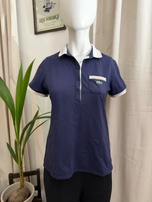 Lacoste Marinblå Pikétröja för dam - Snygg marinblå pikétopp från Lacoste med vit krage och detaljer i vitt och ljusrosa. Toppen har korta ärmar, knappslå framtill och en bröstficka med Lacoste-logga. Perfekt för en sportig och stilren look.