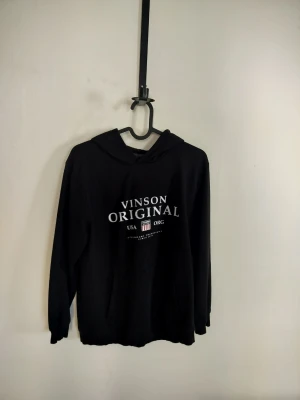 Svart hoodie med tryck Vinson Original - Svart hoodie med vit text 'VINSON ORIGINAL' och USA-detaljer på bröstet. Klassisk modell med huva och känguruficka framtill. Tillverkad i mjukt material och har ribbade muddar vid ärmar och nederkant. Perfekt för en avslappnad och stilren look.