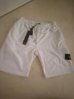Snygga vit Stone Island shorts  - Snygga vita shorts med elastisk midja och snörning framtill. Shortsen har en praktisk sidficka med en svart patch med broderad logga. Perfekta för sommaren och tillverkade i mjuk bomull för maximal komfort. Size L