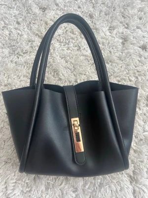 Svart handväska med gulddetalj - Stilren svart handväska i slätt konstläder med två handtag och snygg guldfärgad vridspänne framtill. Väskan har en modern, mjuk form och passar perfekt till dig som gillar minimalistisk design.