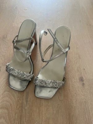 Silvriga pumps med glitterdetaljer - Säljer ett par silvriga pumps med fyrkantig tå och glittrande rem över tårna. Skorna har smala remmar över foten och en hög klack. Perfekta för dig som vill sticka ut med lite extra bling. Materialet är syntet med glitterdetaljer.