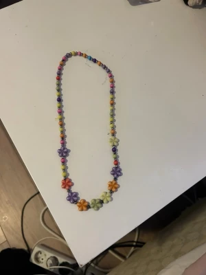 Färgglatt halsband med blommor och pärlor - Säljer ett färgglatt halsband med små runda pärlor och blommor i olika färger. Halsbandet har en lekfull och ungdomlig stil, perfekt för att piffa upp din outfit. 