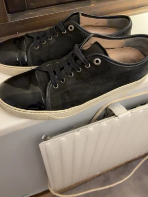 Svarta sneakers med lackdetaljer - Snygga svarta sneakers med ovandel i mocka och glansiga lackdetaljer på tån. Skorna har svarta skosnören, metallfärgade öljetter och vit sula. Perfekta för dig som vill ha en stilren men ändå unik look.