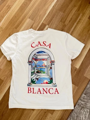 Vit Casablanca t-shirt med tryck - Säljer en vit t-shirt från Casablanca med stort färgglatt tryck på ryggen och mindre tryck på bröstet. Motivet visar en klassisk båge med utsikt över landskap och hav, samt texten 'CASA BLANCA' i rött. T-shirten har rund hals och korta ärmar.