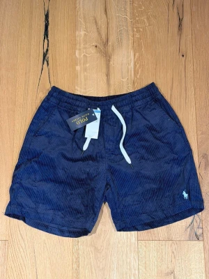 Marinblå manchester shorts Polo Ralph Lauren - Snygga marinblå shorts i manchester från Polo Ralph Lauren med vit snörning i midjan och klassisk broderad logga på benet. Perfekta för sommaren och ger en stilren look med sin ribbade struktur.
