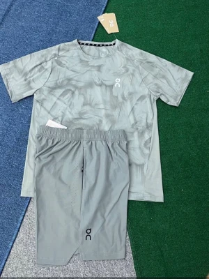 Grått träningsset från On - Snyggt grått träningsset från On med t-shirt och shorts. T-shirten har ett diskret mönster och logga på bröstet, medan shortsen har elastisk midja och logga på benet. Perfekt för löpning eller gympass.
