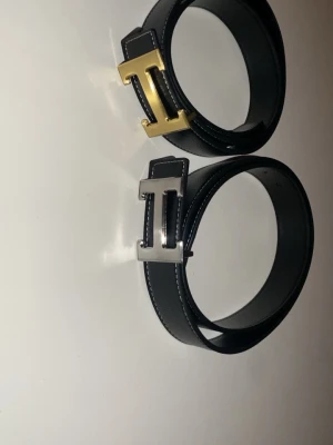 Svarta bälten med H-spänne - Två stilrena svarta bälten med kontrastsömmar och stort H-format spänne, ett i guld och ett i silver. Perfekta för att lyfta vilken outfit som helst och passar till både jeans och kostymbyxor. Gjorda i skinn för en klassisk look.
