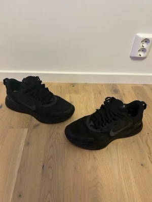 Svarta Nike sneakers med mesh och gummi - Snygga helsvarta Nike sneakers med lågt skaft. Ovandelen är i mesh och syntetmaterial för bra andningsförmåga och komfort. Tjock sula i gummi ger bra dämpning och stabilitet. Perfekta för en sportig och stilren look.