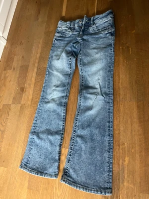 Blå bootcut jeans med utsvängda ben - Säljer ett par klassiska blå bootcut jeans med utsvängda ben och femficksmodell. Jeansen har normal passform och är tillverkade i mjuk bomull med snygga slitningar framtill. Perfekta för dig som gillar retrovibbar och vill ha ett par jeans som sticker ut.