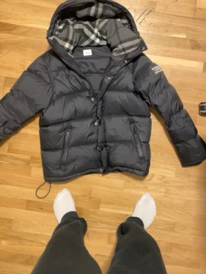 Svart pufferjacka med huva - Snygg svart pufferjacka med avtagbar huva och rutigt foder. Jackan har dragkedja och knappar framtill, två dragkedjefickor och patch på ärmen. Perfekt för kalla dagar och riktigt skön att bära. Materialet är glansigt och tjockt för extra värme.