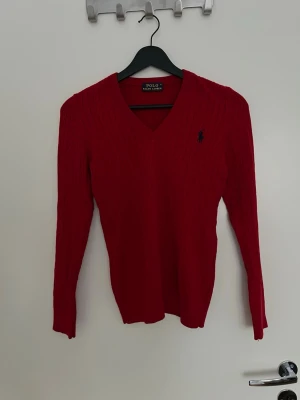 Röd kabelstickad tröja Polo Ralph Lauren - Snygg röd kabelstickad tröja från Polo Ralph Lauren med V-ringning och lång ärm. Tröjan har den klassiska broderade loggan i marinblått på bröstet och är tillverkad i mjukt material som sitter tajt och bekvämt. Perfekt för lager på lager och stilrena outfits. Inte använd.