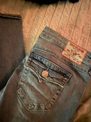 Ljusblå True Religion jeans - Säljer ett par ljusblå jeans från True Religion med ikonisk bakficka och tydliga sömmar. Klassisk femficksmodell med knappdetalj och patch med logga bak. Perfekta för en avslappnad och trendig look.