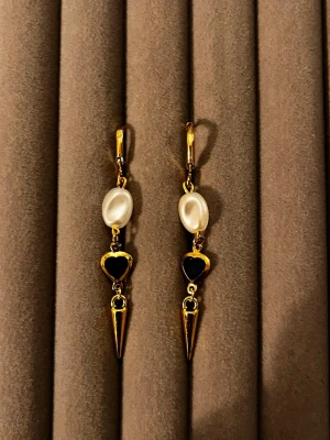 Guldörhängen med pärlor och hjärta - Säljer ett par unika guldtonade örhängen med vita pärlor, svarta hjärtan i metall och spetsiga hängen. Örhängena har en cool och edgy design som sticker ut. Perfekta för dig som vill ha något annorlunda och trendigt i smyckeskrinet.