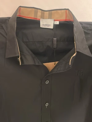 Svart skjorta från Burberry - Stilren svart skjorta från Burberry med klassisk krage och knäppning framtill. Skjortan är helt ny och aldrig använd. 