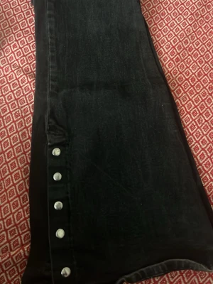 Svarta jeans med knappar längs benet - Säljer ett par svarta jeans med raka ben och coola silverfärgade knappar längs ena sidan. Jeansen har klassisk femficksmodell och hög midja. Materialet är denim i bomull och de har en snygg tvättad look.