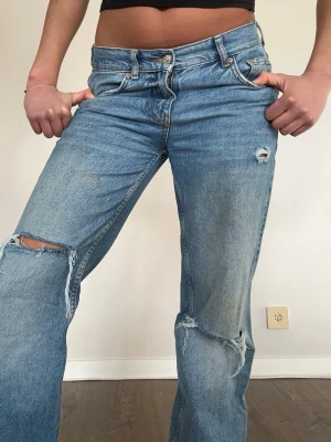 Blå slitna raka jeans - Ljusblå raka jeans med slitningar och hål på knäna. Jeansen har klassisk femficksmodell, med rise midja och knappgylf. Perfekta för en avslappnad och trendig look.