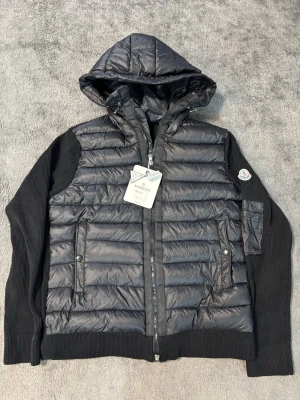 Moncler cardigan - Snygg svart cardigan från Moncler med stickade ärmar och huva. Jackan har två framfickor med tryckknappar, dragkedja framtill och Moncler-logga på ärmen. Kroppen är quiltad och glansig medan ärmarna är ribbstickade för en cool kontrast.