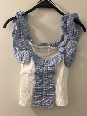 Vit och blå rutig volangtop från Zara - Supersöt topp från Zara med vita ribbade partier och blå/vit rutigt mönster på volangprydda axelband och framtill. Toppen är ärmlös och har knappar framtill samt volangdetaljer som ger en romantisk vibe. Perfekt för dig som gillar unika detaljer och trendig stil.