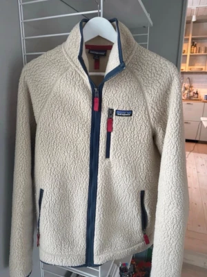 Beige fleecejacka från Patagonia - Mysig beige fleecejacka från Patagonia med blå detaljer och dragkedja framtill. Jackan har hög krage, tre fickor med dragkedja och långa ärmar. Perfekt för kyliga dagar och outdoor vibes.