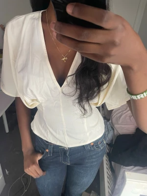 Vit omlottopp med knytband - Säljer en vit omlottopp med v-ringning och vida, korta ärmar. Toppen har ett snyggt knytband i ryggen som markerar midjan och ger en feminin siluett. Perfekt att styla med jeans eller kjol för en trendig look.