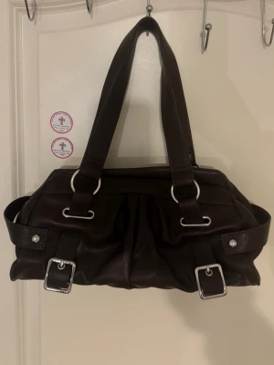 Mörkbrun handväska med spännen - Snygg mörkbrun handväska i fejkläder med dubbla axelband och silverfärgade metalldetaljer. Väskan har dekorativa spännen framtill och veckad design som ger en cool look. Perfekt storlek för det viktigaste och passar till många outfits.