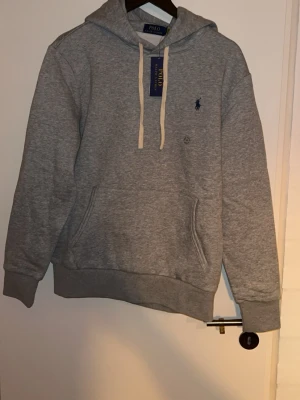 Grå hoodie från Polo Ralph Lauren - Snygg grå hoodie från Polo Ralph Lauren med klassisk huva och vita dragsnören. Tröjan har känguruficka framtill och den ikoniska lilla loggan broderad på bröstet. Tillverkad i mjukt bomullsmaterial som är perfekt för en avslappnad stil. Hejsan alla! Jag har precis fyllt 18 år och har fått en massa presenter. Det är mycket av presenterna som inte passar min stil, men det är väldigt fina och de som bär dessa kläderna har fin stil. Det är därför jag väljer att sälja detta.  Allt är oanvänt