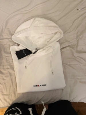 Saint Laurent hoodie - Säljer en clean vit hoodie från Saint Laurent med broderad logga i svart och rött framtill. Hoodien har huva med dragsko och en stor magficka. Skön passform och riktigt stilren look, perfekt till jeans eller sweatpants. 100% bomull jag är 178 väger 57