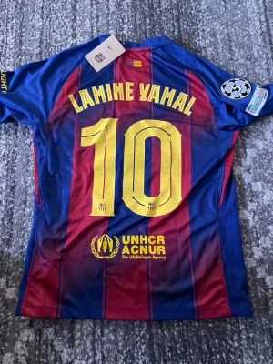FC Barcelona matchtröja Lamine Yamal - Säljer en officiell FC Barcelona matchtröja från Nike med Lamine Yamal och nummer 10 på ryggen. Tröjan har klassiska blå och röda ränder, klubbmärke, Nike-logga och Spotify som sponsor framtill. Tillverkad i lätt och ventilerande material, perfekt för fotbollsträning eller match.