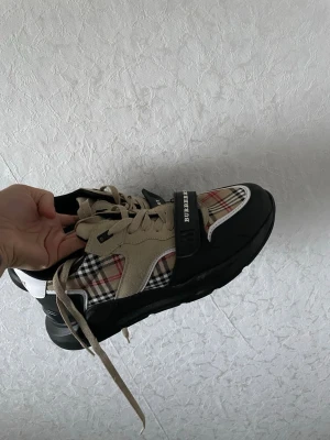 Burberry sneakers med rutigt mönster - Snygga sneakers från Burberry med klassiskt beige, svart och rött rutmönster, svarta detaljer och beige mockaliknande partier. Skorna har snörning och en svart rem med Burberry-logga över tån. Perfekta för dig som vill sticka ut med stilrena detaljer.