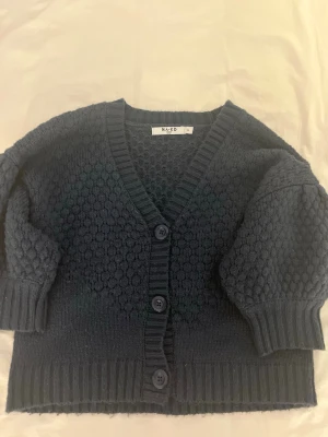 Mörkblå stickad kofta från NA-KD 💙 - Mysig mörkblå stickad kofta från NA-KD med v-ringning och tre knappar framtill. Koftan har ballongärmar och ett strukturerat mönster över hela plagget. Ribbstickade muddar vid ärmslut och nederkant ger en snygg finish.💙🦋