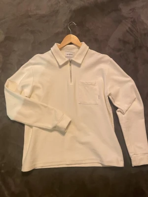 Vit halvzip tröja från Woodbird - Snygg vit långärmad tröja från Woodbird med halvzip och krage. Tillverkad i ekologisk bomull, har en bröstficka på vänster sida och en clean, minimalistisk look. Perfekt för dig som gillar stilrena plagg med en modern touch.