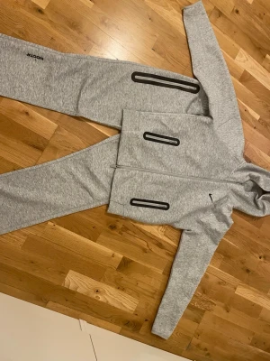 Gråa mjukisbyxor från Nike x Nocta - Säljer ett par gråa mjukisbyxor från Nike x Nocta med svart logga och coola svarta detaljer längs benen. Byxorna har dragsko i midjan och raka ben. Perfekta för chill dagar eller när du vill ha en sportig look.