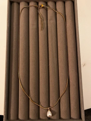 Guldhalsband med pärlhänge - Snyggt och stilrent halsband i guldton med en droppformad vit pärla som hänge. Kedjan är tunn och har ett justerbart lås med förlängningskedja. Perfekt för dig som gillar minimalistiska smycken med en elegant touch.