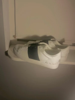 Valentino opens  - Snygga vita sneakers med en bred svart rem över sidan. Klassisk låg modell med vita snören och sula. Tillverkade i slätt läderliknande material som ger en clean och modern look. Perfekta för dig som gillar stilrena och trendiga skor.