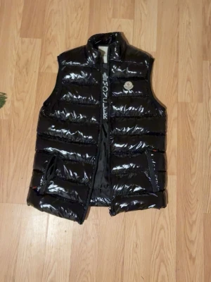 Moncler väst - Snygg svart dunväst från Moncler med glansigt material och hög krage. Västen har två framfickor med dragkedja och Moncler-logga på bröstet. Köpte den när modellen kom ut helt ny för 8000 har tyvär ej kvar kvittot