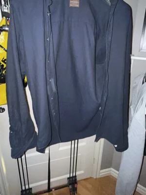 Mörkblå skjorta med knappar - Stilren mörkblå skjorta från Oscar Jacobson med klassisk krage och knäppning framtill. Skjortan har lång ärm och är tillverkad i ett mjukt bomullsmaterial. Perfekt för en clean och enkel look.