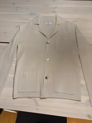 Beige cardigan från Bläck - Stilren beige cardigan från Bläck med tre knappar framtill och tre praktiska fickor. Tröjan har klassisk krage och är tillverkad i ett mjukt, stickat material. Perfekt för en clean och avslappnad look. Perfekt för lite finare tillfällen! Pris går att diskutera vid snabb affär! Hör gärna av er!