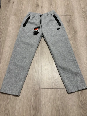 Gråa mjukisbyxor från Nike - Snygga grå mjukisbyxor från Nike med svarta dragkedjor på fickorna och justerbar midja med snörning. Byxorna har en rak passform och är tillverkade i mjukt bomullsmaterial. Perfekta för chill dagar eller träning.