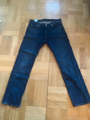 Mörkblå raka jeans från Levi's - Säljer ett par klassiska mörkblå jeans från Levi's med rak passform. Jeansen har tydliga sömmar, knappgylf och är tillverkade i slitstark denim. Perfekta för en avslappnad och tidlös stil. 