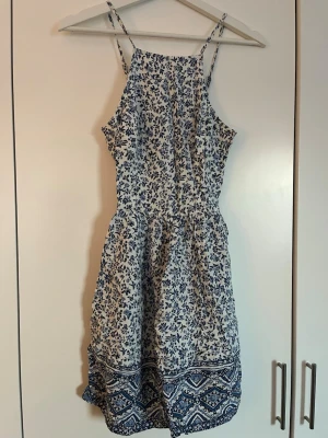 Blåvit blommig sommarklänning - Söt blåvit klänning med smala axelband och blommigt mönster. Klänningen har en kort längd och avslutas med en dekorativ bård i blått och vitt, samt knytning i ryggen för perfekt passform. Perfekt för varma dagar och har en lätt, luftig känsla, trots dubbelt bomullstyg. Har ett litet hål i tyget längt upp på kanten (försökt visa på sista bilden), men inget som syns på eller något som lätt blir större.