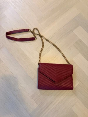 Vinröd kuvertväska med kedjerem - Snygg vinröd kuvertväska i quiltat konstläder med guldfärgad kedjerem och v-format mönster. Väskan har ett stilrent kuvertlock och axelrem i kombinerat kedje- och lädermaterial. Perfekt accessoar för att lyfta din outfit.