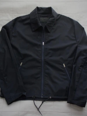 Prada Windbreaker - Stilren mörkblå Prada windbreaker med dragkedja framtill, två snedställda fickor med dragkedja och justerbar dragsko i nederkant. Klassisk krage och raka ärmar. Perfekt för blåsiga dagar och enkel att matcha till olika outfits. Alla dragkedjor har ingraverade loggor, även triangeln och labeln placerad på framsidan. Den har även snören för att kunna spänna/ lossa den. NFC funkar. Hör av er vid minsta intresse! 