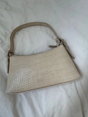 Beige axelremsväska från NA-KD - Snygg beige handväska från NA-KD i croco-mönstrad syntet med axelrem. Väskan har dragkedja upptill och flera innerfack för smart förvaring. Perfekt till din vardagsstil och rymmer det viktigaste du behöver ha med dig.